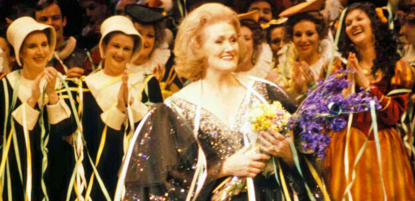 Dame Joan Sutherland on stage holding flowers with the cast applauding her.