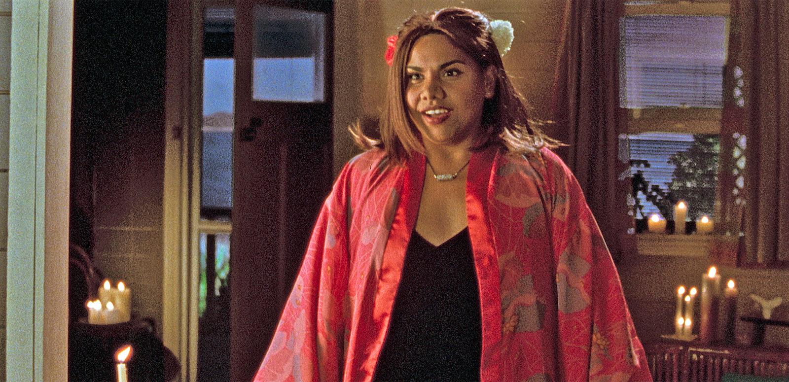 Deborah Mailman in Radiance A young Aboriginal woman wearing a red kimono and flower in her hair stands in the middle of a candlelit room facing camera with a half-smile on her face.