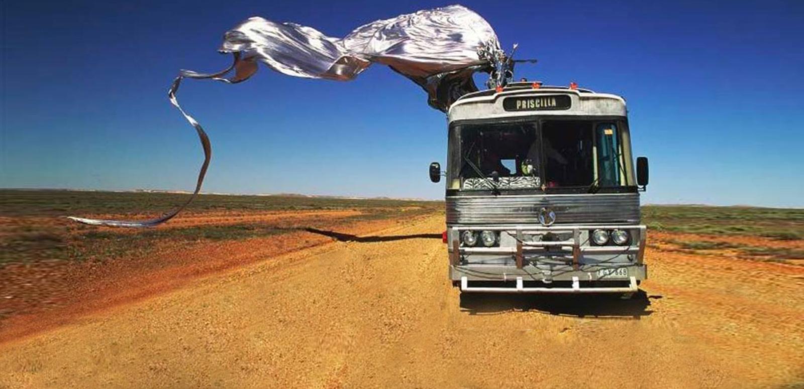 Priscilla Queen of the Desert 25th Anniversary Scene from Priscilla, Queen of the Desert. A tour bus on a dirt road in the desert with blue sky in the background. On top of the bus is Guy Pearce in a shiny silver frock sitting on a giant stiletto shoe with a long train flowing behind the bus.