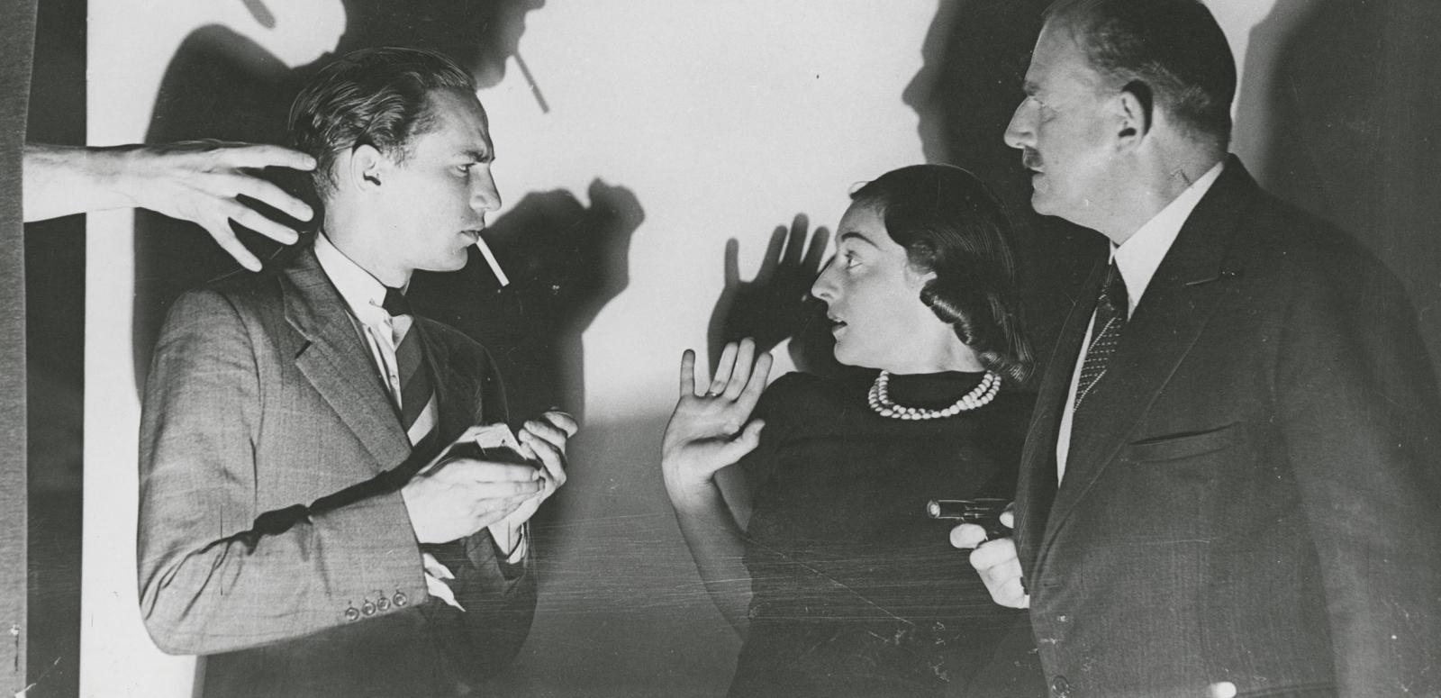 Two men and a woman. One man lights a cigarette while a mysterious hand reaches for him. The woman looks terrified. The other man holds a gun.