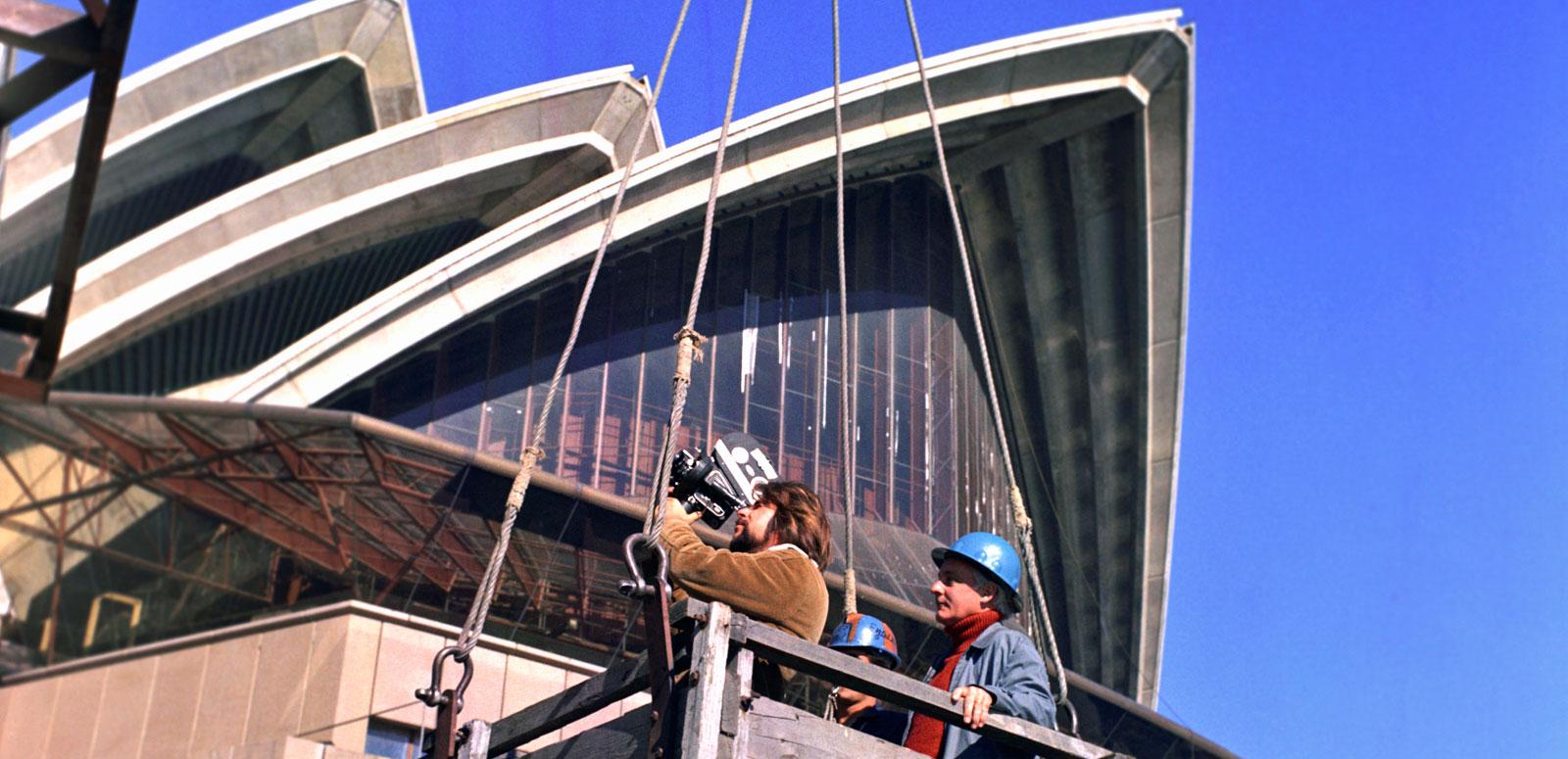 Newscaf: Sydney Opera House The sails of the Opera House tower above a camera operator and a man in a hardhat. They are standing in a wooden box which is hoisted to a crane.