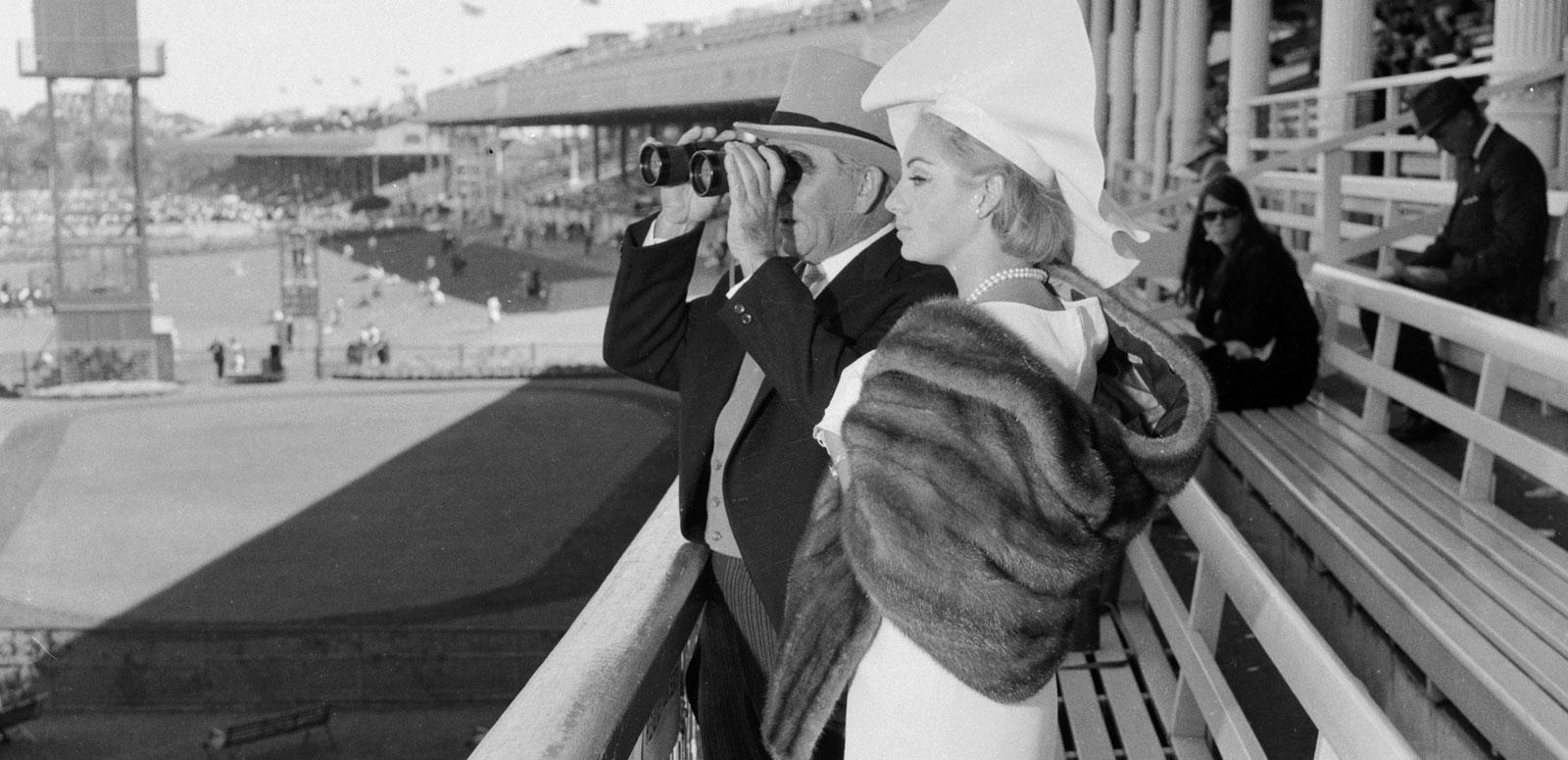 Melbourne Cup Woman's Day ad latest A man and woman are dressed for a day at the races. He is looking through binoculars. Black and white image.