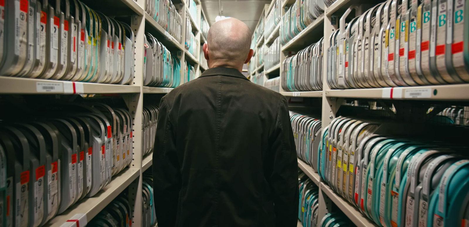 A person has their back to the camera and is standing amidst shelves that are lined with video tapes.