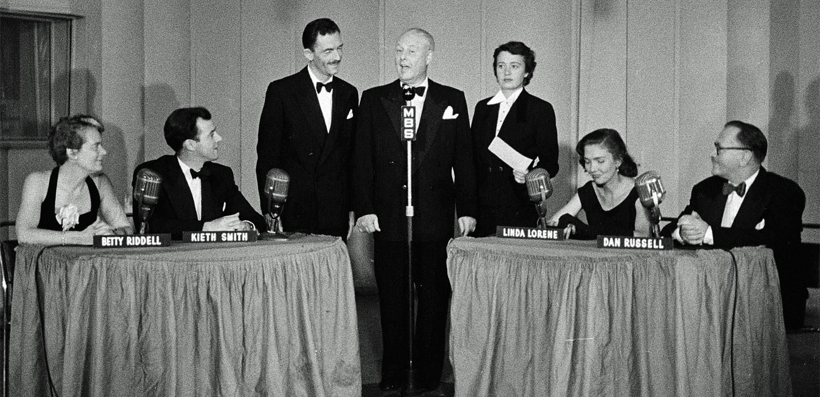 A studio with 3 people standing in the centre in front of a radio microphone and a table on each side with two panellists seated at each table.