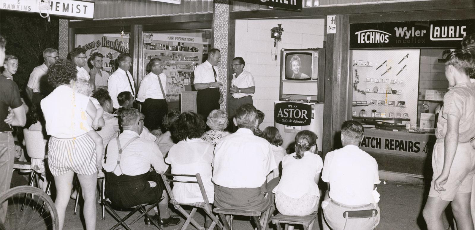 Crowd watching test TV transmission in Townsville A crowd of people (some seated and some standing). They are watching a television set through a shop window.