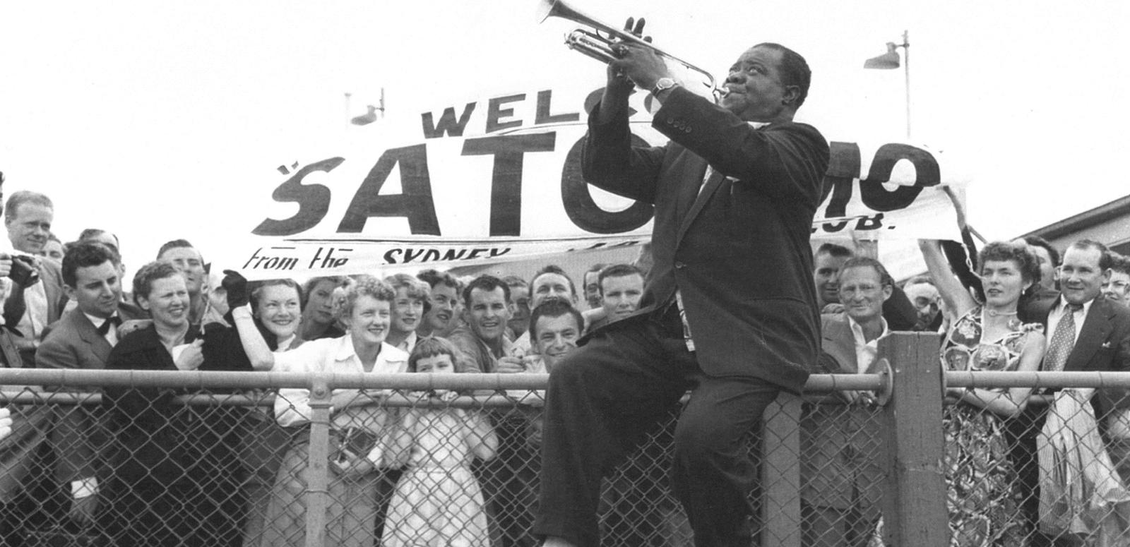 Louis Armstrong at Sydney airport in 1956, He's sitting on the top rail of a wire fence, playing the trumple to a crow gathered there to welcome him.