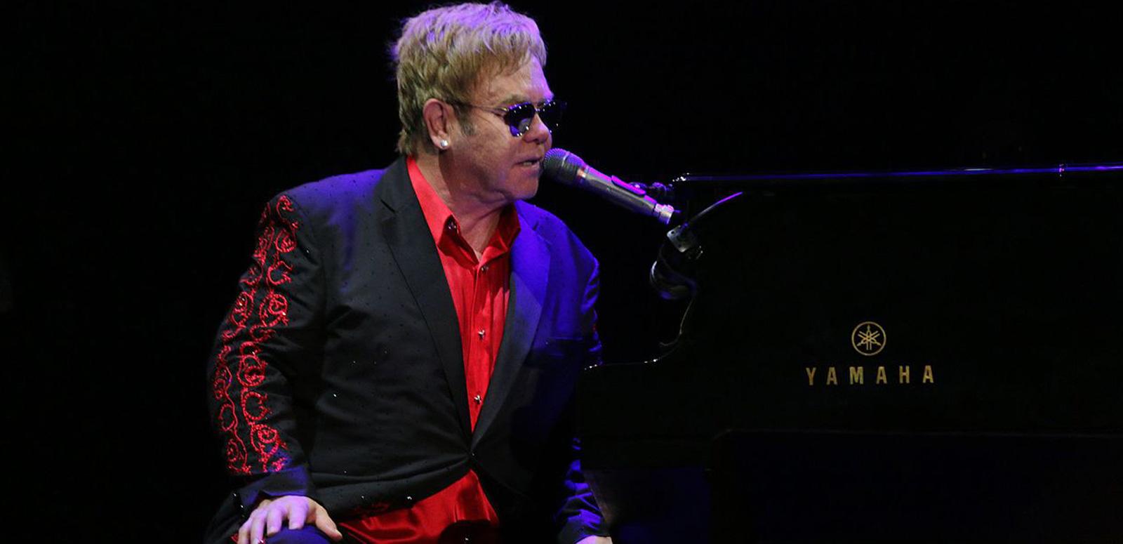 Musician Elton John sitting at a piano and singing into a microphone. He is wearing a black and red suit and dark glasses. 