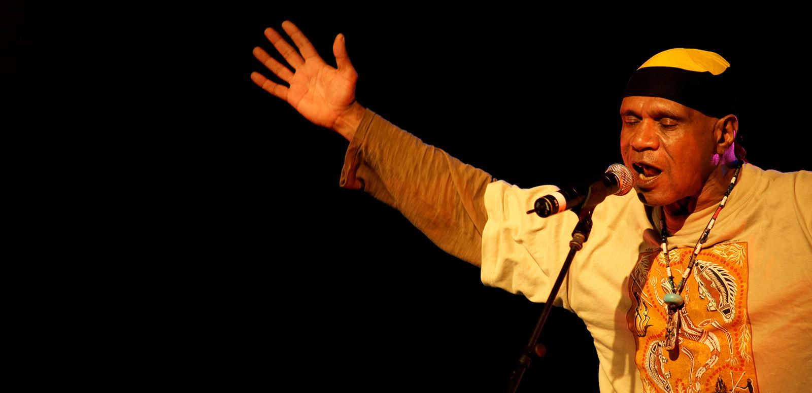 Archie Roach in concert Archie Roach stands at a microphone singing. He is holding his arms outstretched.