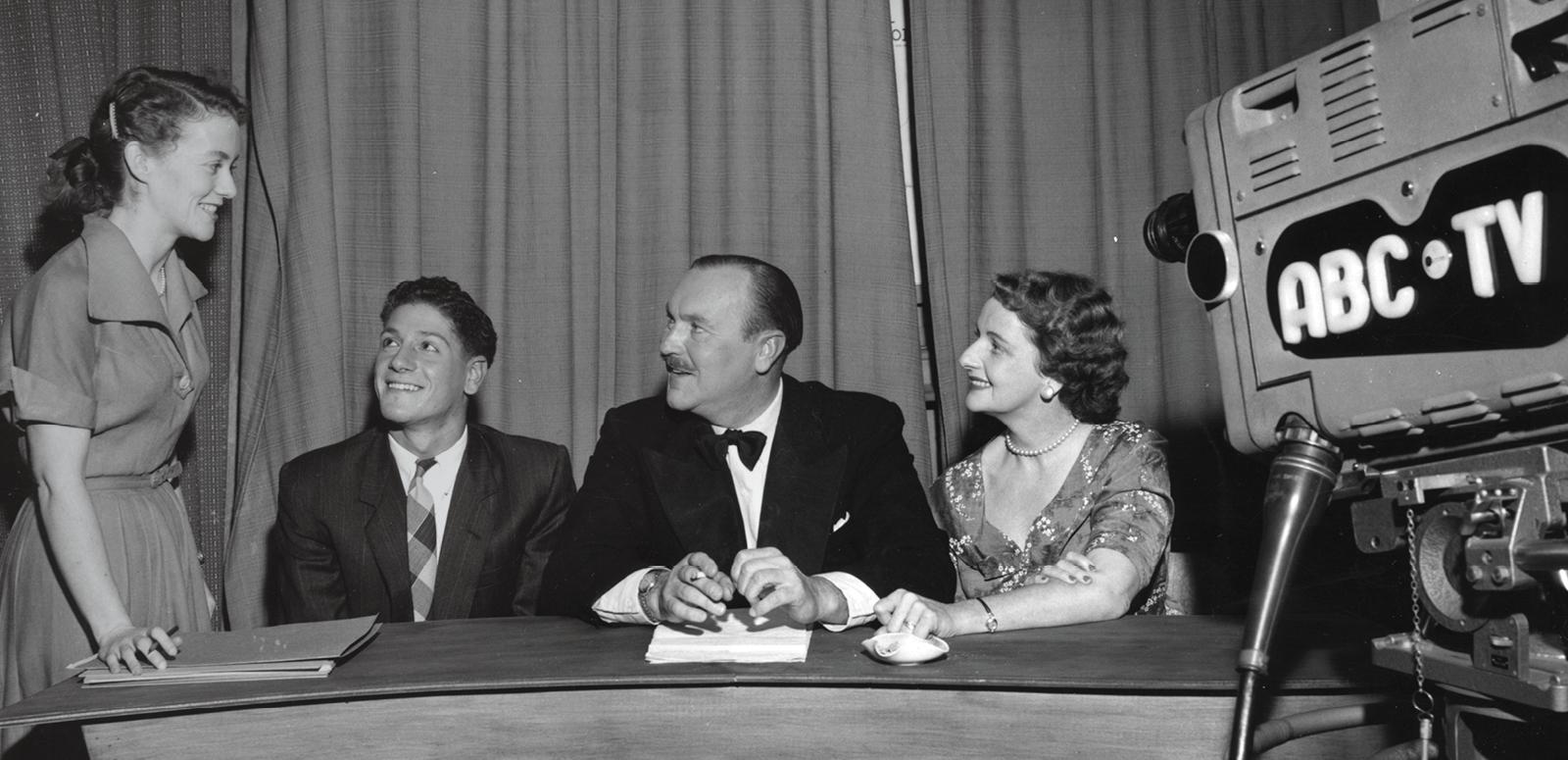 Four people are in a studio setting. Three of them sit behind a desk and the fourth stands. They are all looking at her. There is a film camera pointed at them that says ABC TV on the side.