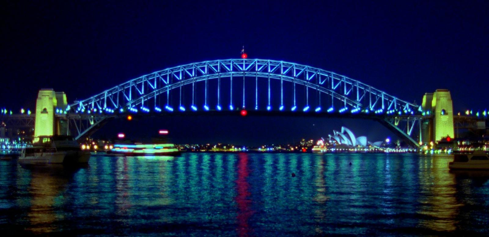 Night - Sydney Harbour Bridge A nightime photo of the Sydney Harbour Bridge showing it all lit up. The bridge arch is blue with red lights at the top and bottom centre. The pylons are lit yellow. The water shows the reflections of the passing ferry and the Sydney Opera House.