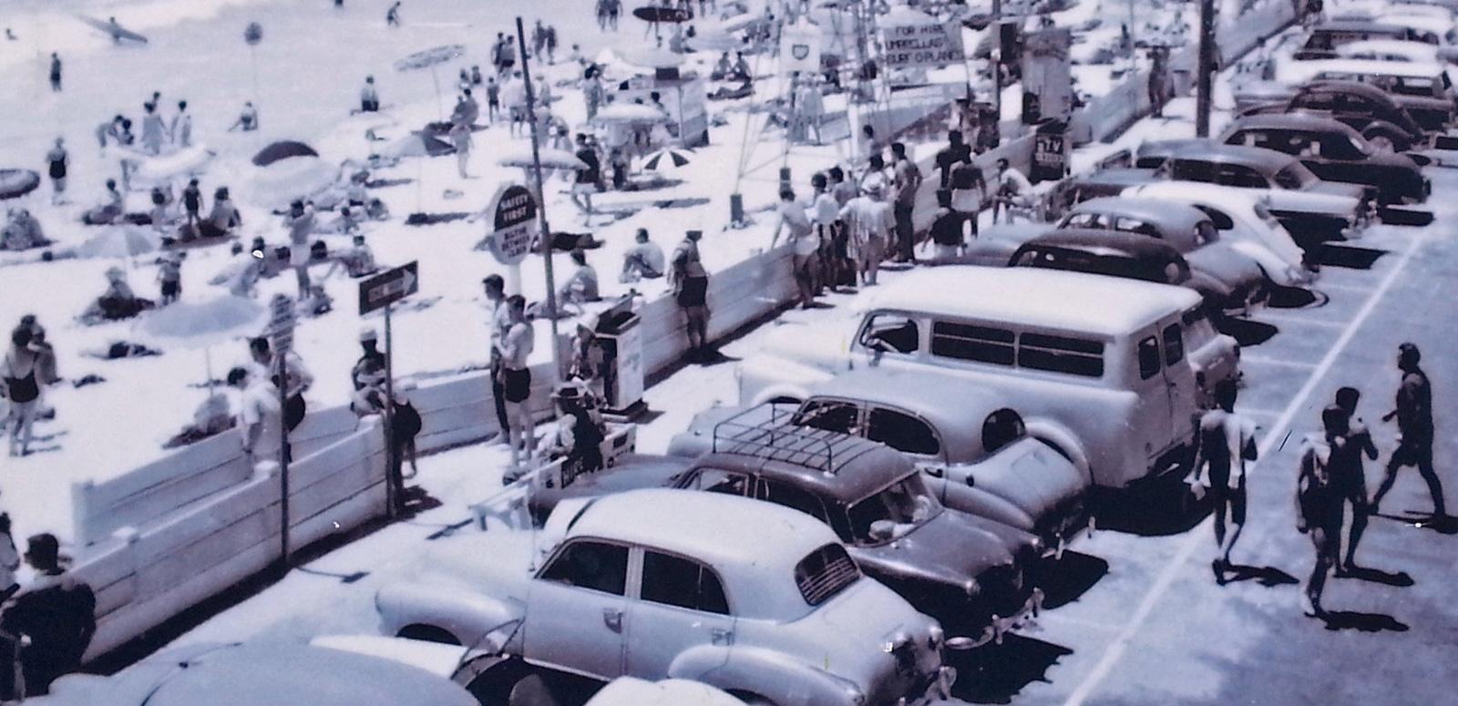 A busy Australian beachfront showing people on the sand and cars behind the sand in a parking lot, circa 1960s.
