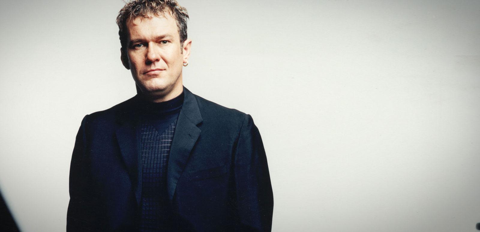 Jimmy Barnes wearing a black suit and shirt and looking directly at camera. Photographed from waist up.