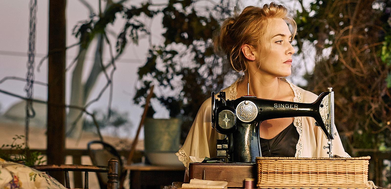 Kate Winslet in a scene from The Dressmaker. She is seated outside on a porch behind a table with a vintage singer sewing machine on top and looking sideways into the distance.