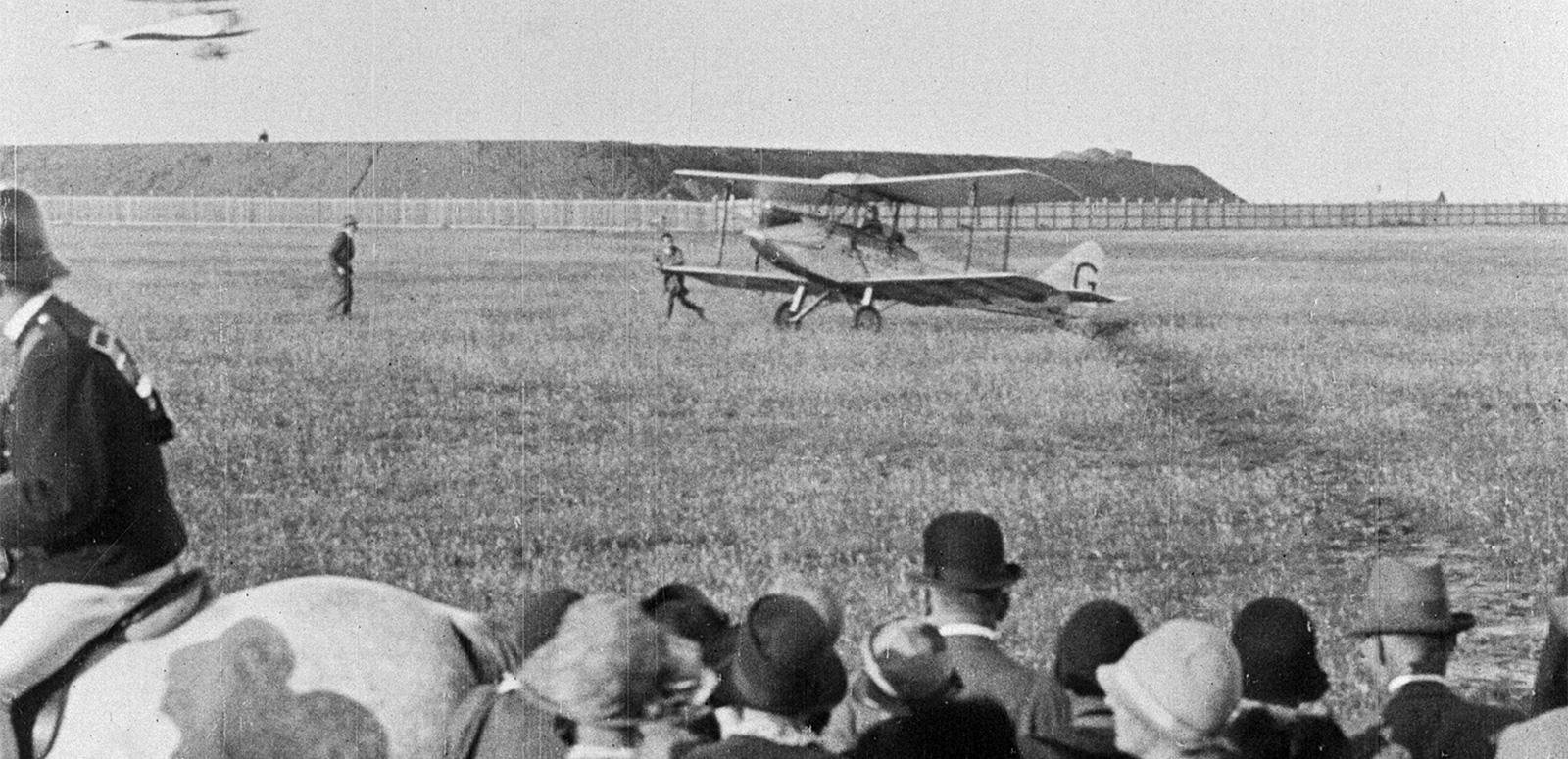 An early 20th century biplane lands in a field, watched by a crowd of spectators An early 20th century biplane lands in a field, watched by a crowd of spectators
