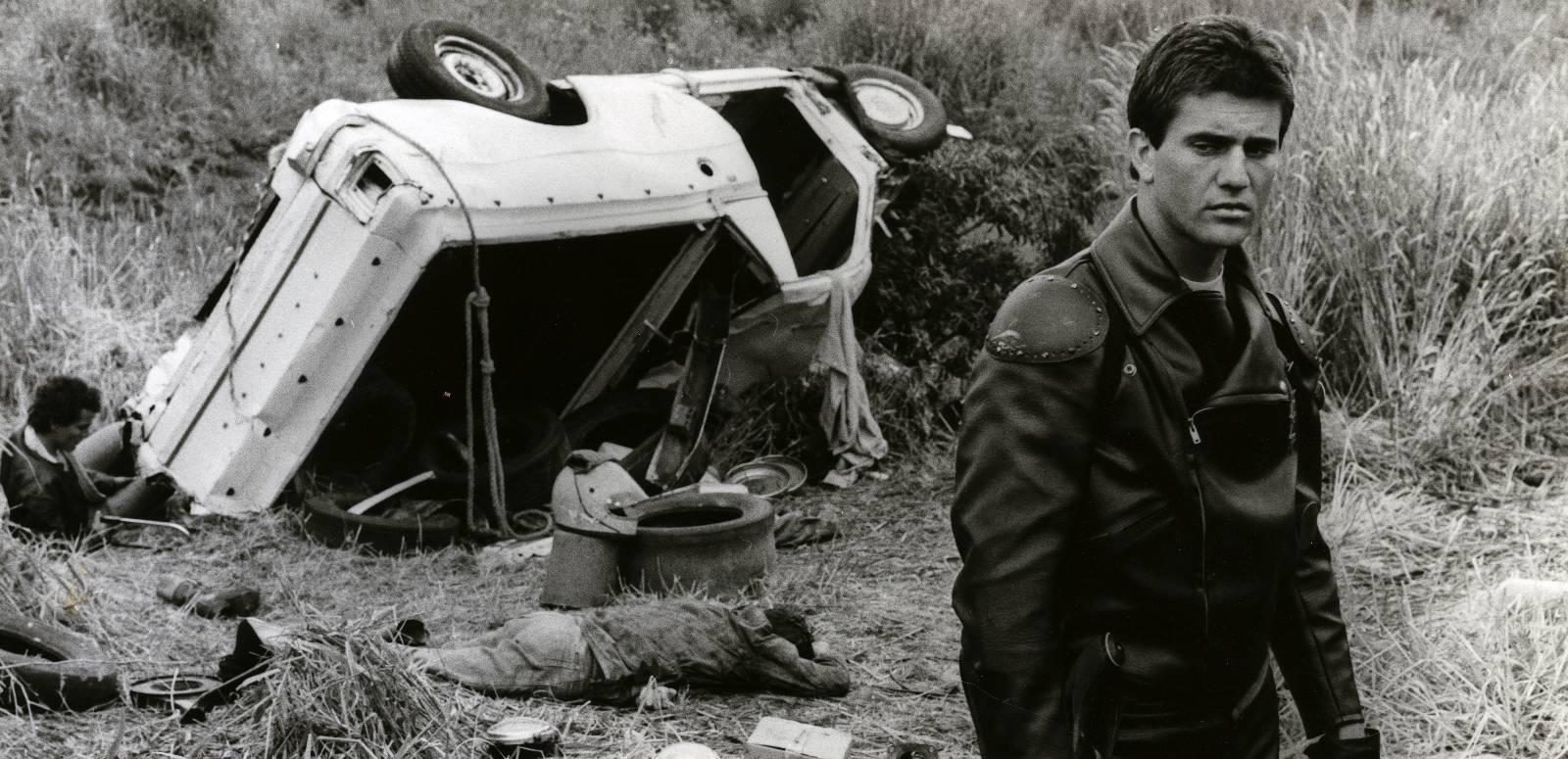 Mad Max dressed in his motorcycle cop leathers stands in front of an overturned car, looking dispassionately away