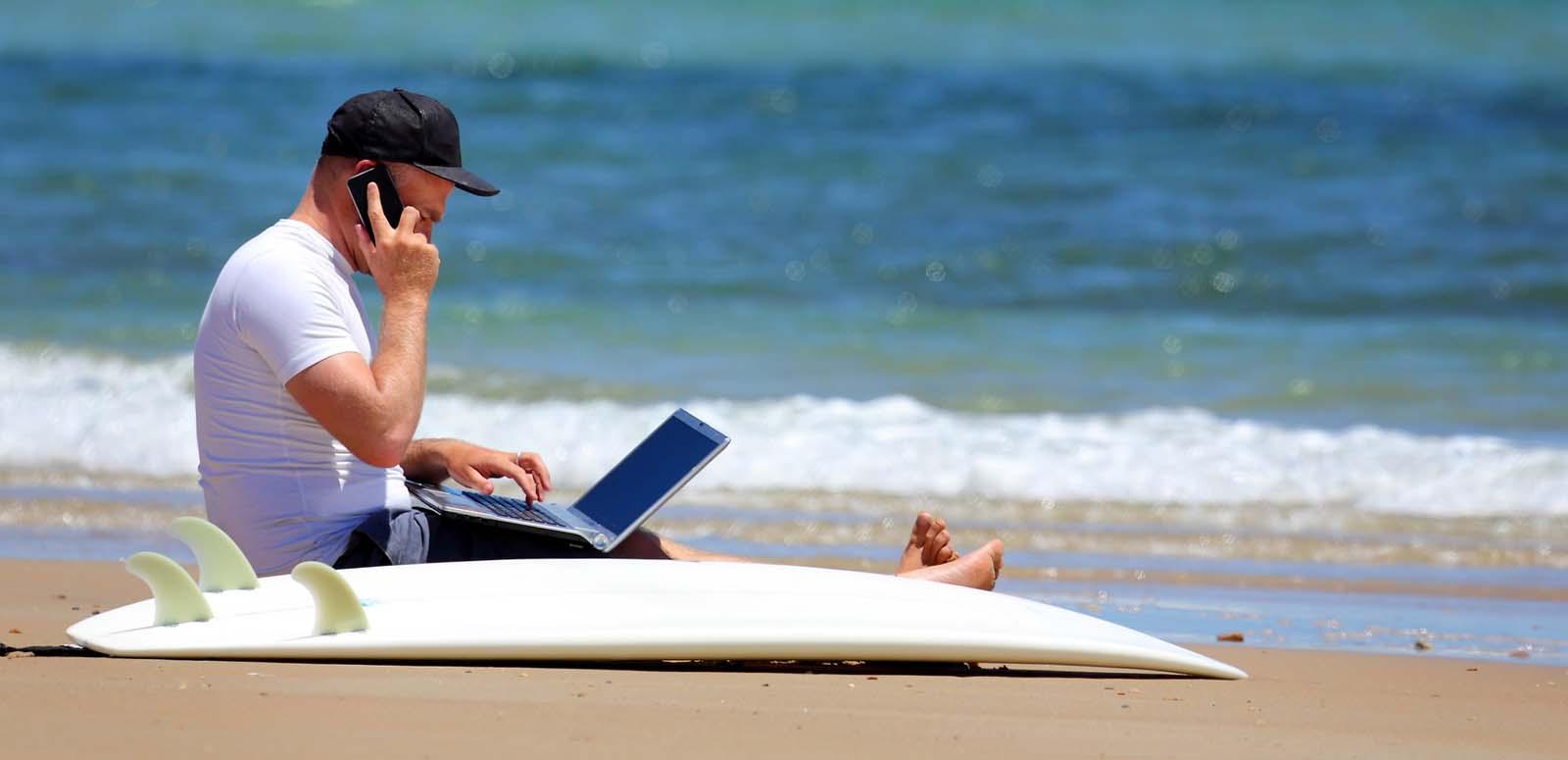 A man working on the beach with a mobile phone and laptop and a surfboard on the sand next to him