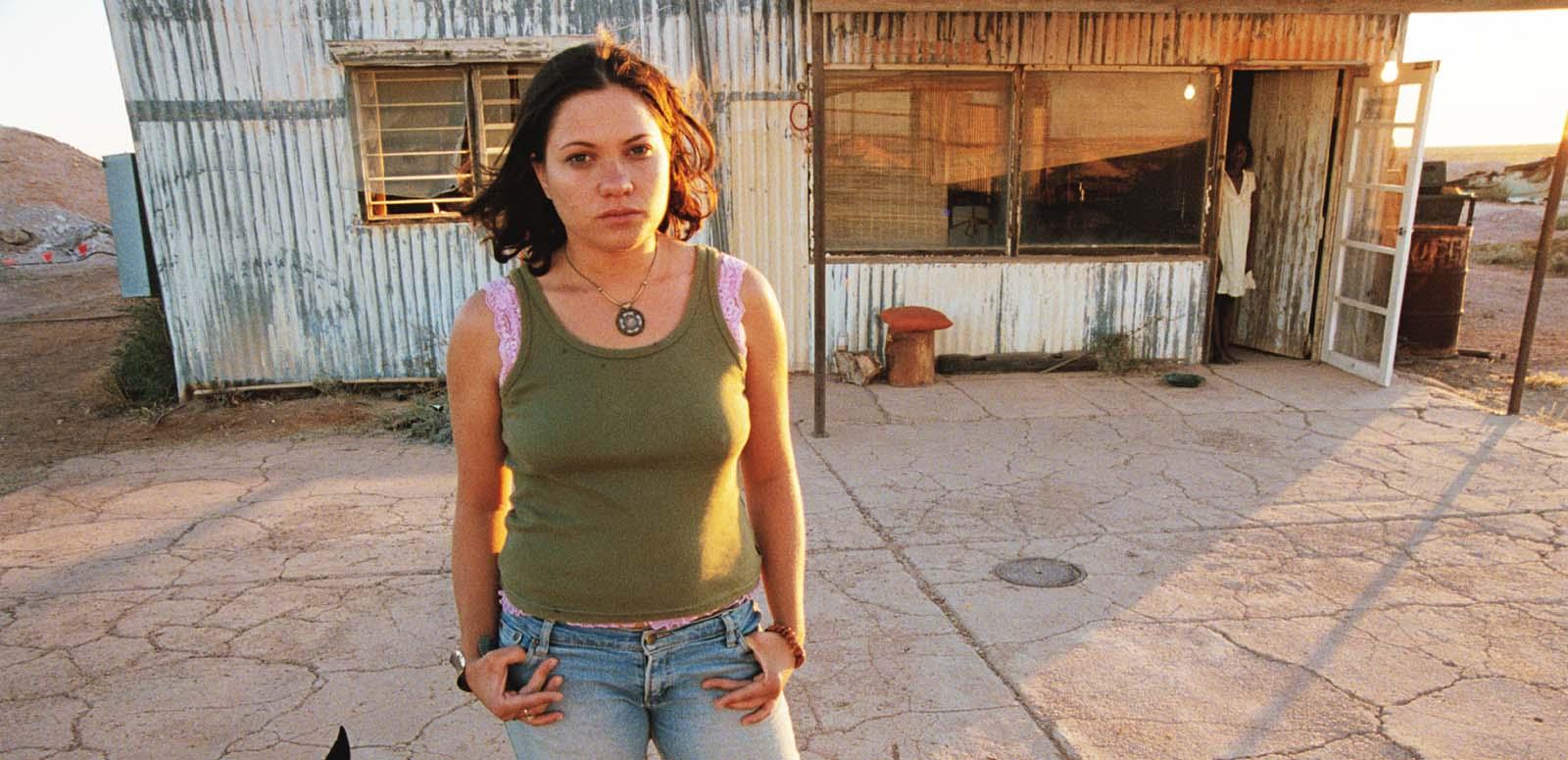 A young woman stands outside a corrugated iron building; an older Aboriginal woman stands in the doorway behind her