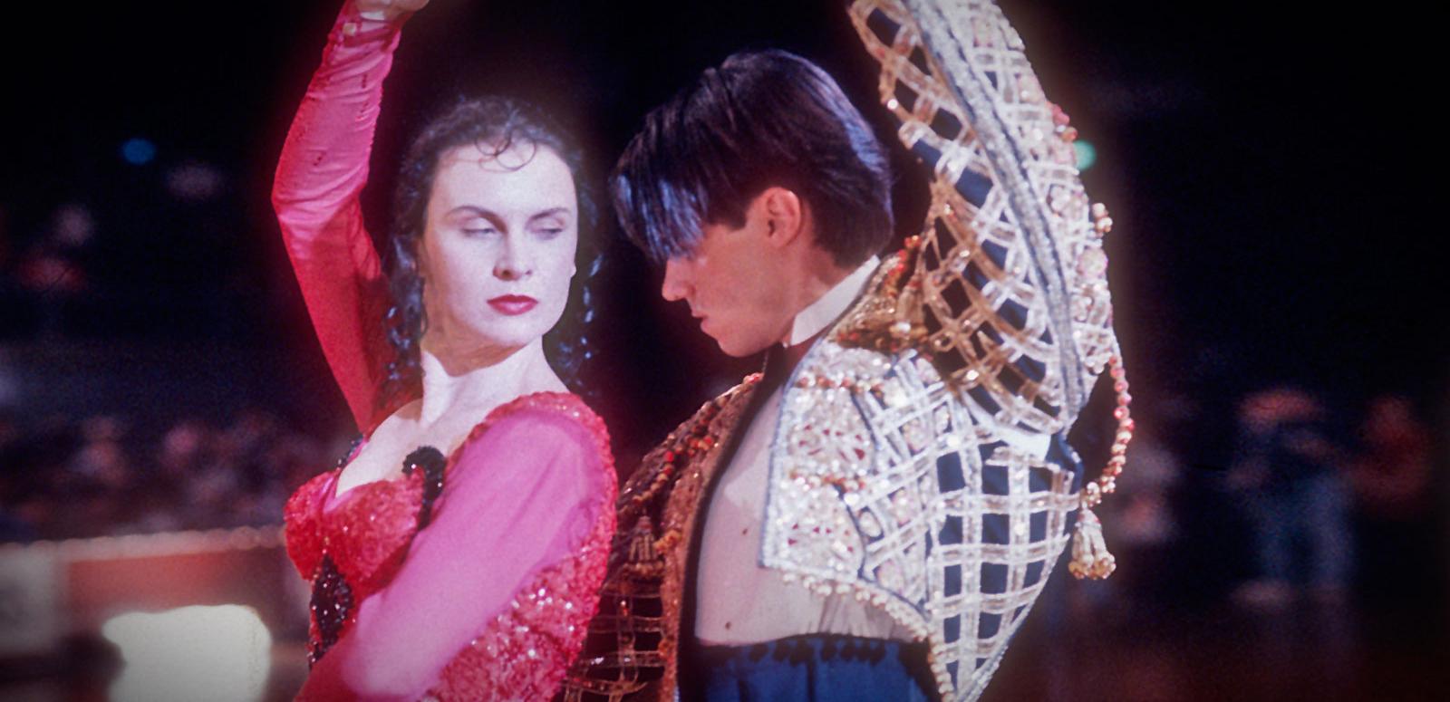 Tara Morice and Paul Mercurio in a dance pose in a scene from the film Strictly Ballroom. She is wearing a red and black beaded and ruffled flamenco style dress and he is wearing a spanish style gold and black bolero jacket.