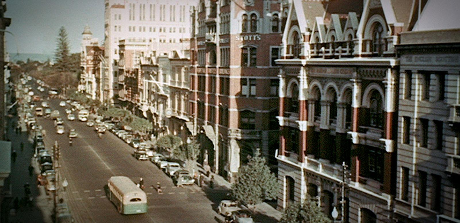 A street scene from Perth circa 1954. A street scene from Perth circa 1954. The street is lined with 3 and 4 story buildings and there are many cars parked on the side of the road, there are trees along the footpaths and a few pedestrians can be seen crossing the street.
