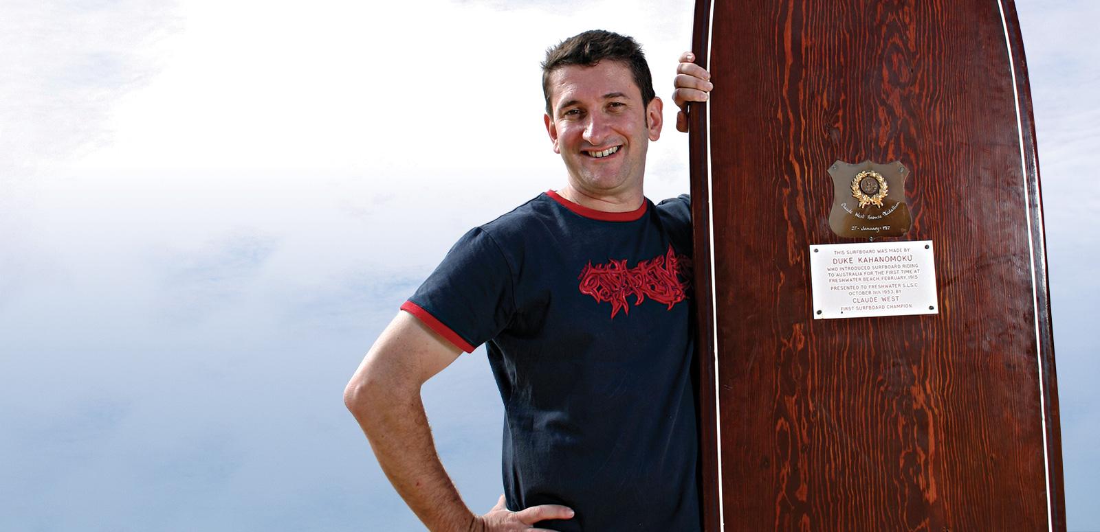 Warren Brown standing next to a large wooden surfboard Warren Brown standing next to a large wooden surfboard