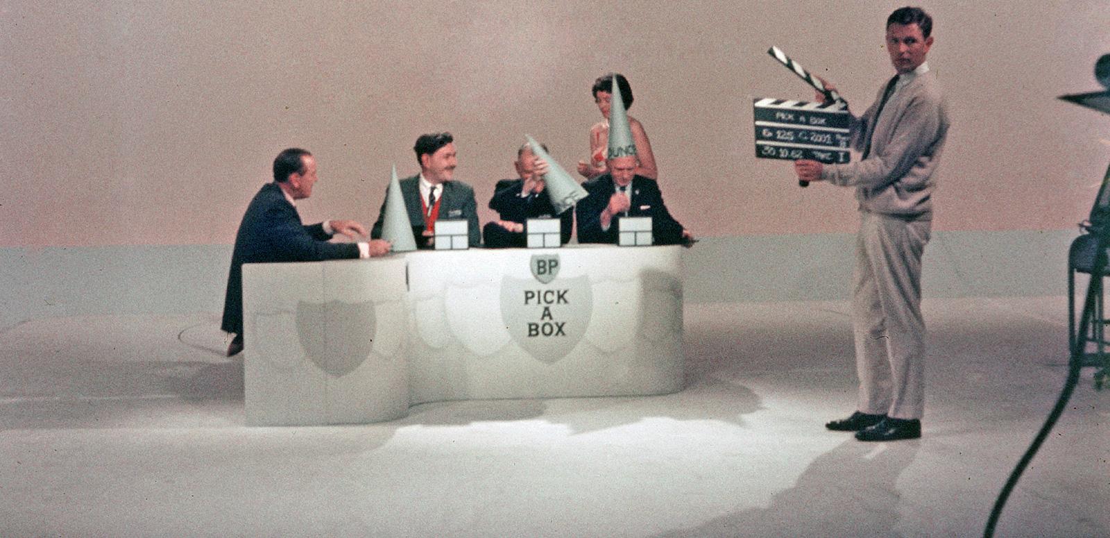 A group of people behind a desk in a TV studio with a man standing in front of them holding a clapper board. 