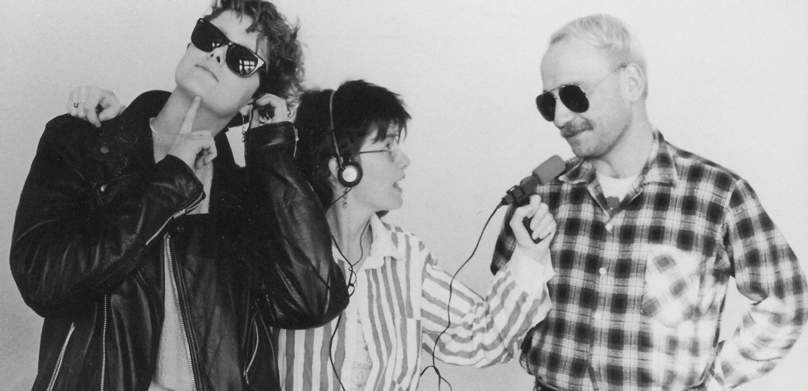 A woman wearing headphones aims a radio microphone on a cord at a man standing next to her. Another person leans against the woman, as if listening in on their recorded conversation.