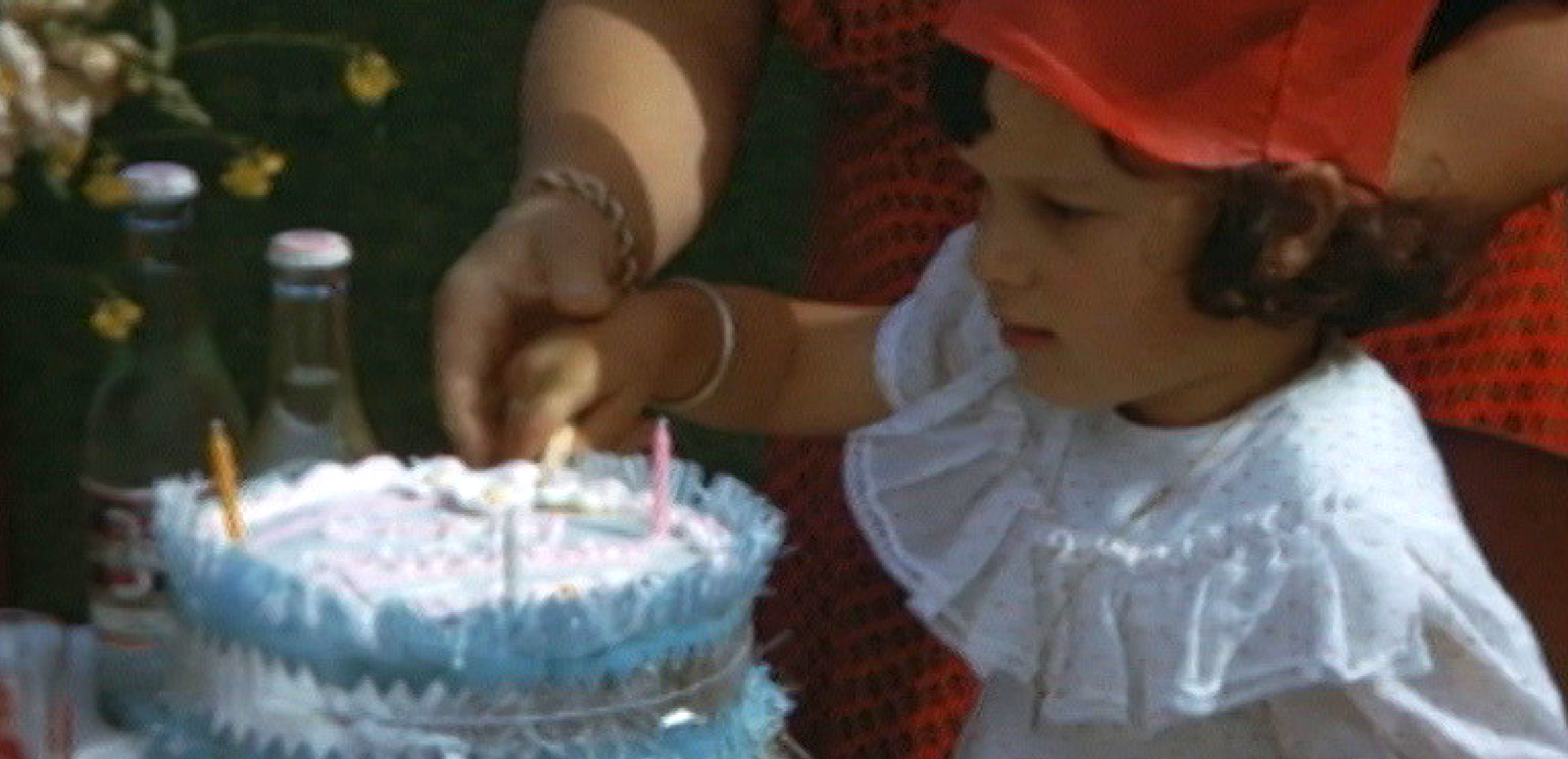 A little girl in a red party hat is looking at her birthday cake with candles on it and blue paper fringing around the outside of the cake.