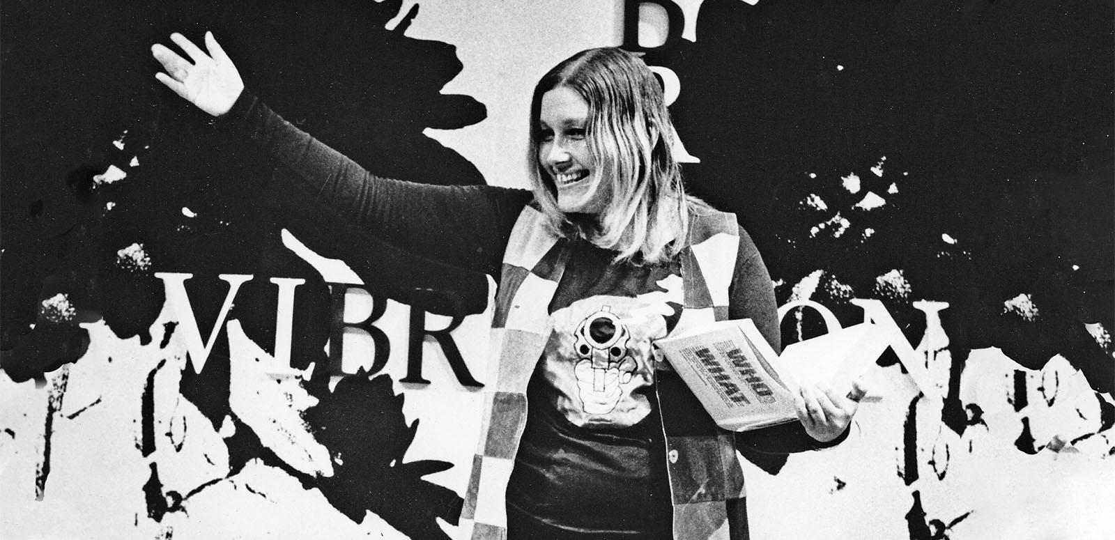 Journalist Lillian Roxon waving at a crowd. She is holding a book.