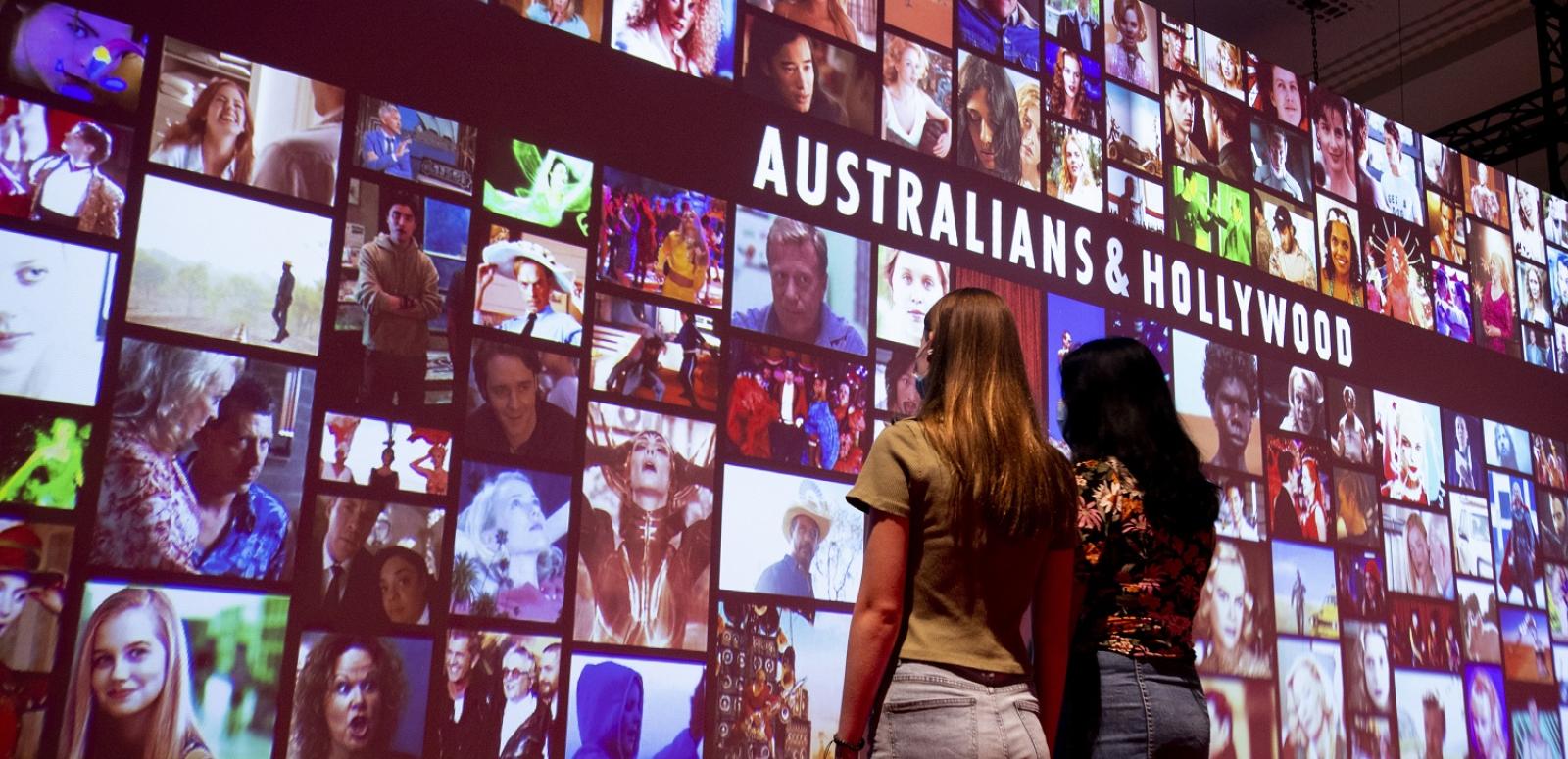 Two young women standing in front of a large screen with the faces from Australian film industry Two young women standing in front of a large screen with the faces from Australian film industry