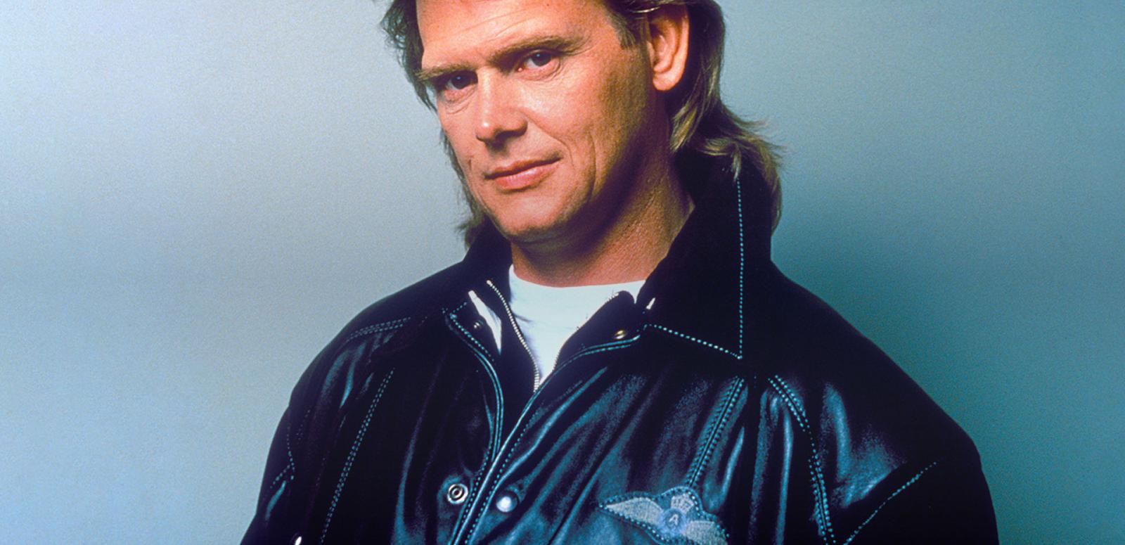 Head and shoulders shot of John Farnham wearing a black leather jacket looking at camera with head slightly tilted.