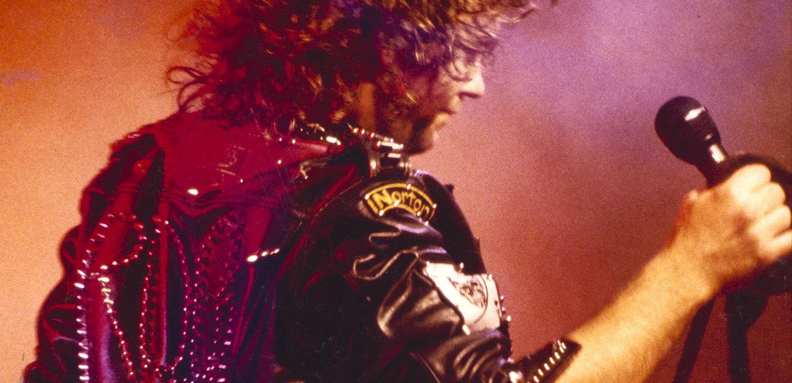 Jimmy Barnes wearing a black leather jacket and holding a microphone. He is pictured from the waist up and turned away from the camera.