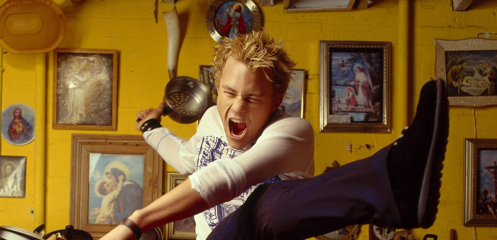 Actor Heath Ledger striking a dramatic pose with his arms out, one leg up in the air and his mouth open wide and eyes shut. He is in a yellow kitchen. Actor Heath Ledger striking a dramatic pose with his arms out, one leg up in the air and his mouth open wide and eyes shut. He is in a yellow kitchen.
