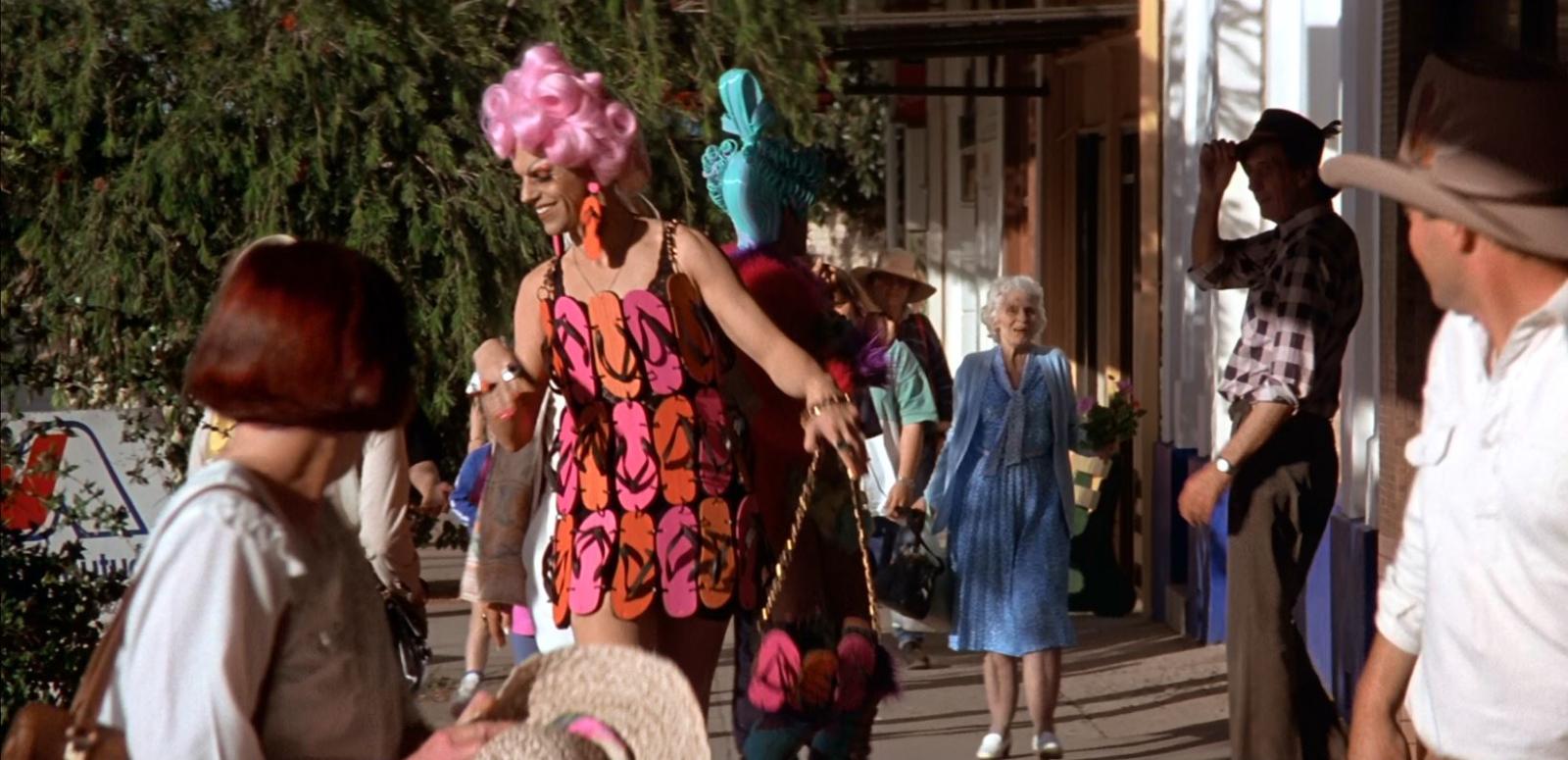 Hugo Weaving with a bright pink wig and wearing a dress made out of pink and orange flip-flops. He is walking down a footpath in drag make-up and attracting surprised looks from passersby