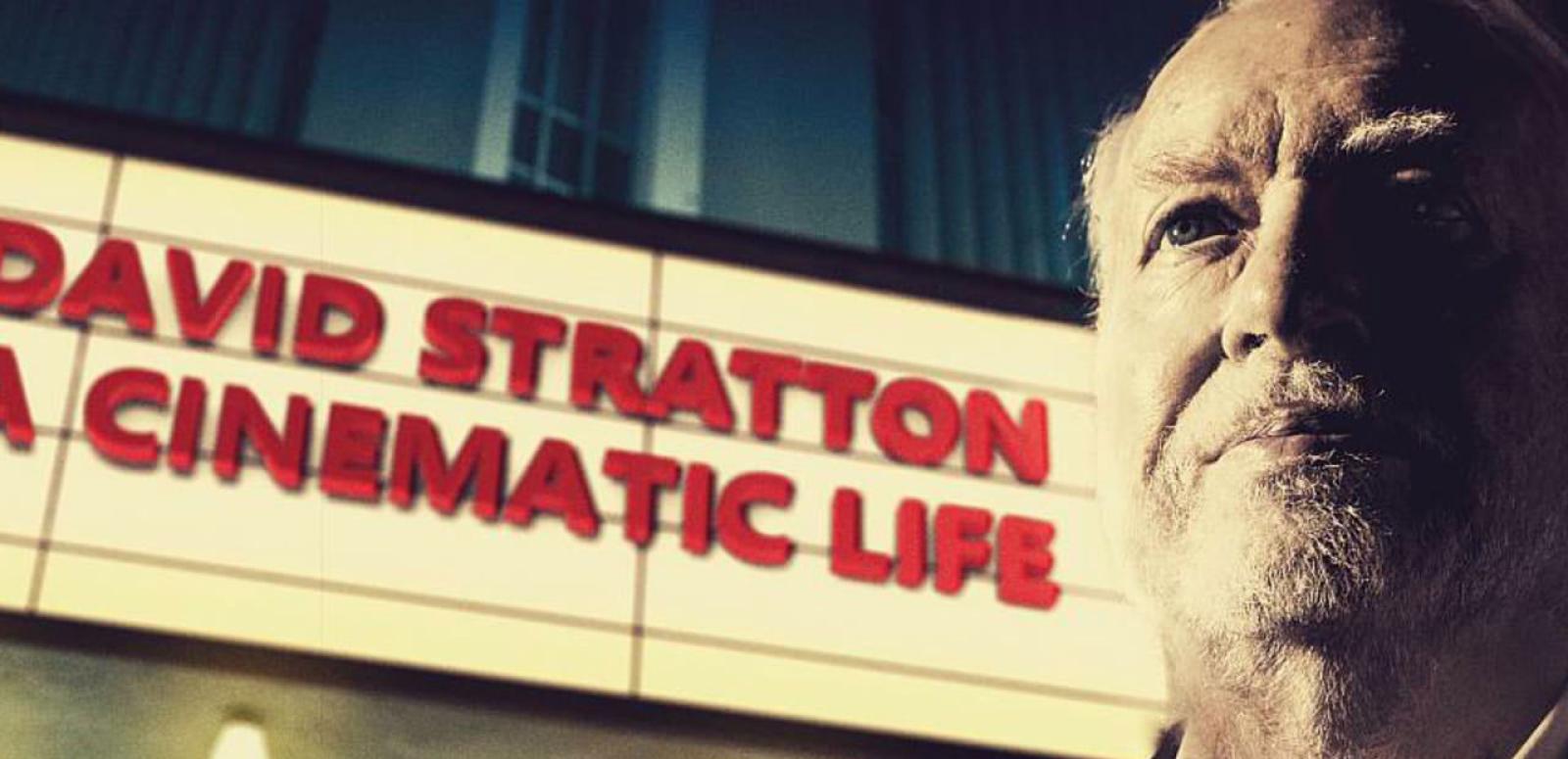 Close up of David Stratton standing next a sign that reads: David Stratton a cinematic life