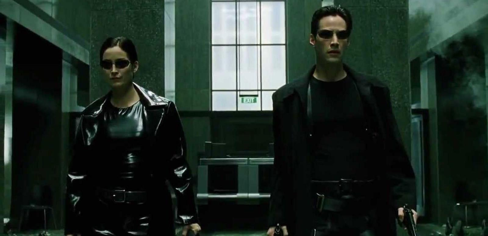 A man and a woman wearing dark sunglasses, black clothes and carrying guns.