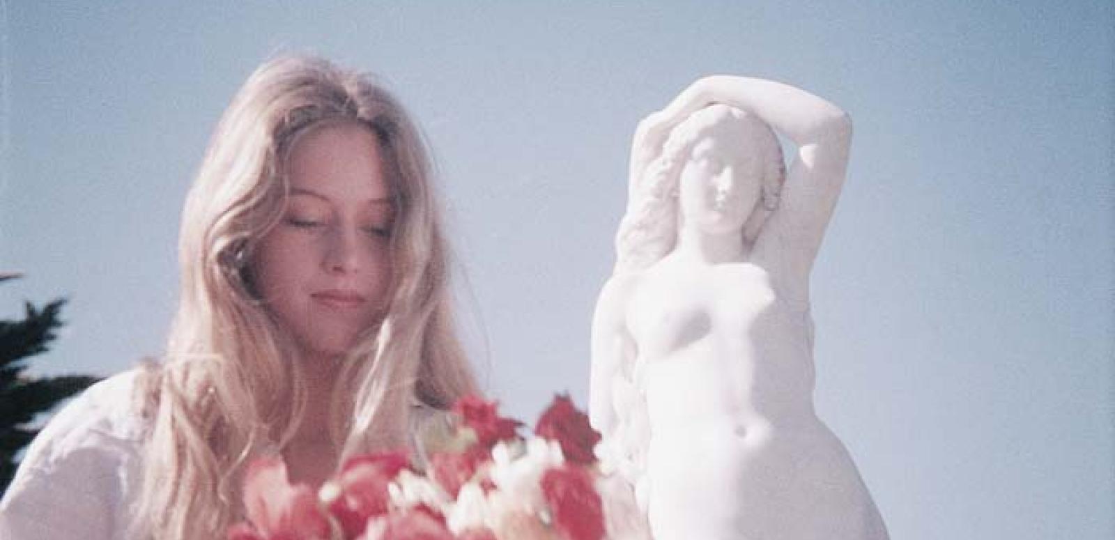 Miranda holds a bouquet of colourful flowers next to a statue of a woman in an outtake from Picnic at Hanging Rock.