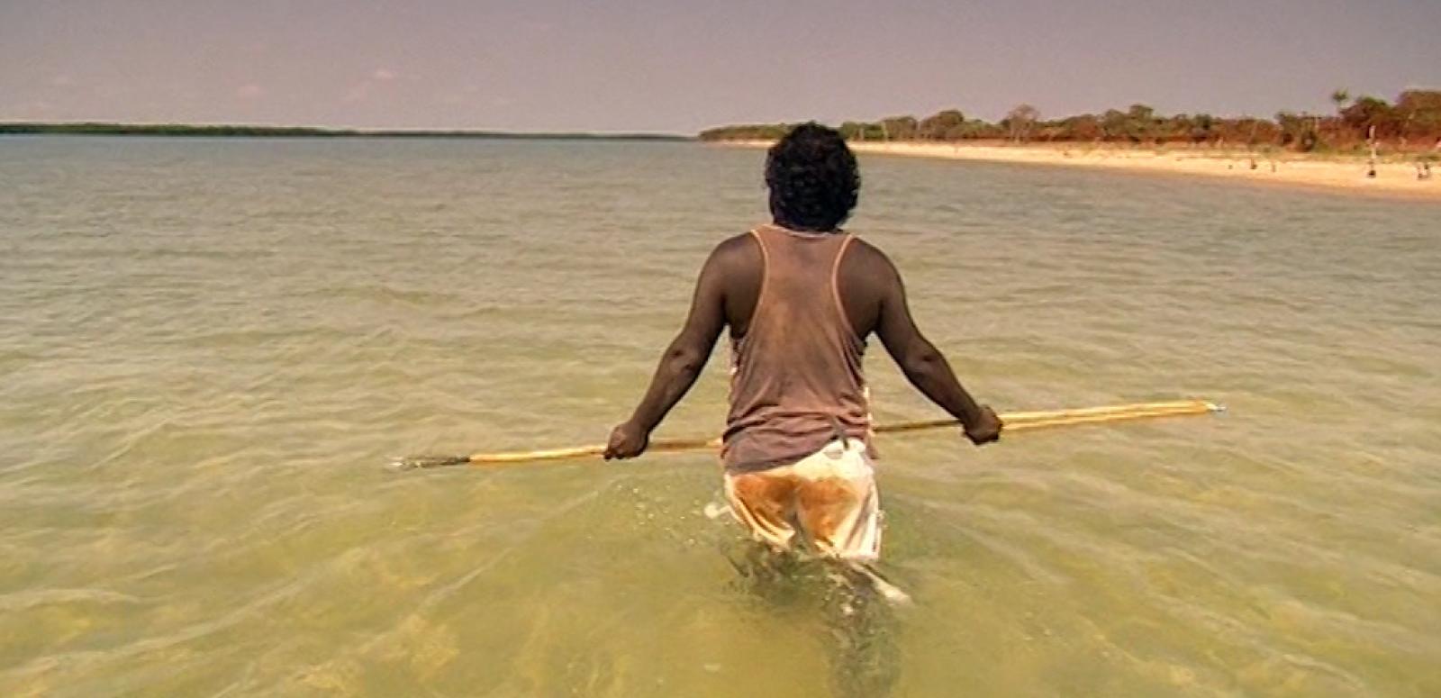 An Indigenous man walking through the water holding a spear