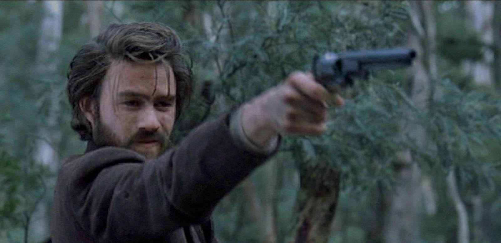 Heath Ledger as Ned Kelly holds a gun out in front of him.