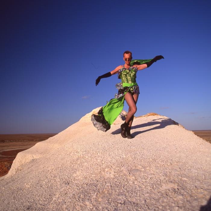 Hugo Weaving in a green dress dancing on a sand dune