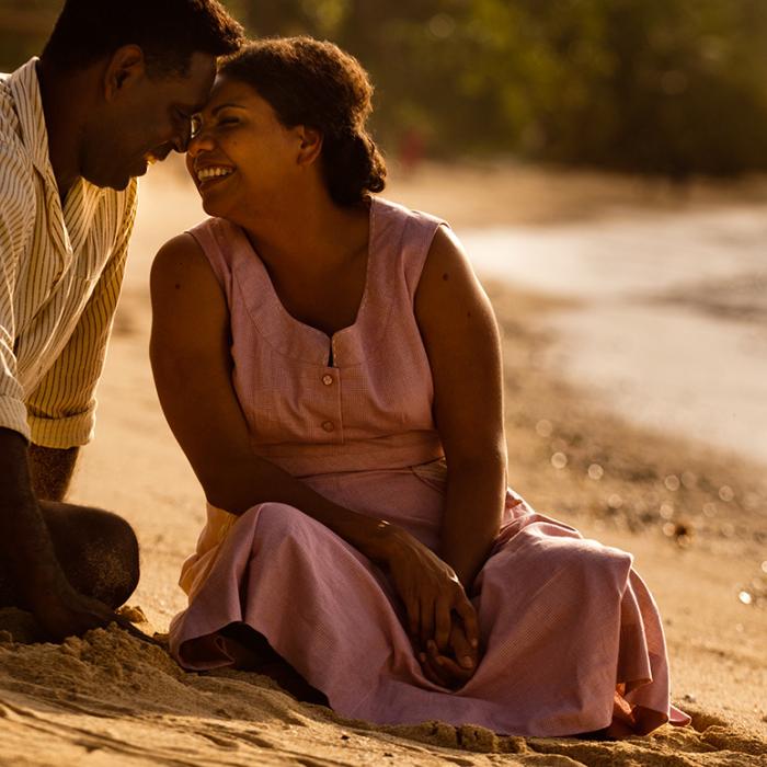 A First Nations man and woman sit together on a beach looking at each other and smiling in a scene from Mabo.
