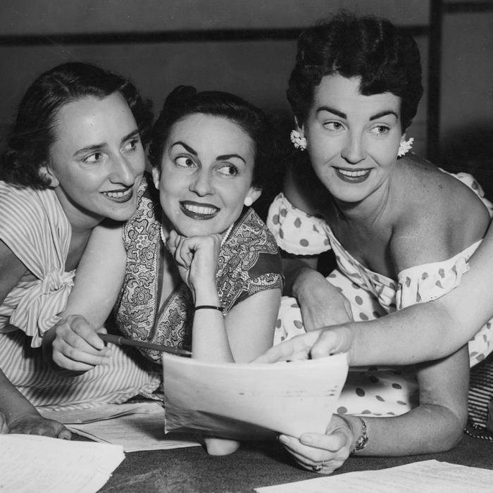 Five women leaning against a desk looking at a sheet of paper, they are all smiling. 