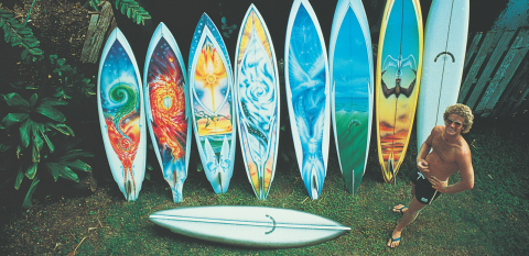 A man in board shorts standing next to 8 colourfully illustrated surfboards leaning against a fence