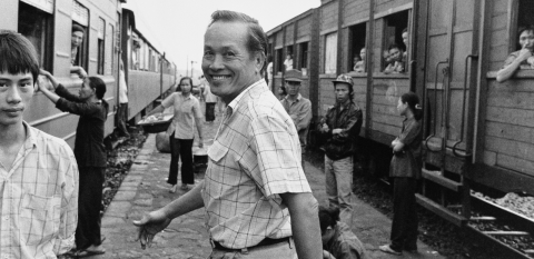 A man smiling and looking backwards as he walks between two stationary trains in Vietnam. Around him are people walking, watching and looking at the camera