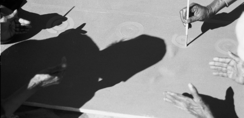 A close-up of women's shadows and hands painting the ancestral design of the Sugar leaf dreaming
