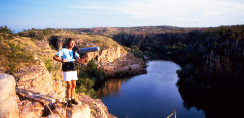 A crew member holding a boom mike stands on a rocky canyon edge above a pool of water