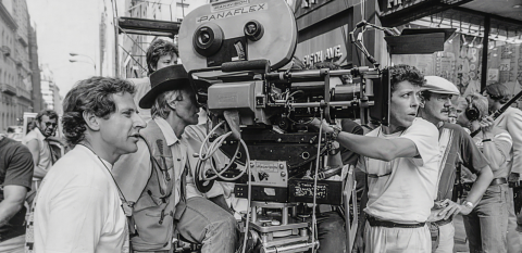 Paul Hogan looks through the viewfinder of a large movie camera while filming Crocodile Dundee on the streets of Manhattan, surrounded by other crew members.