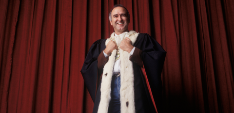 Larry Hand in ceremonial mayoral robe on stage laughing, with red curtains behind him.