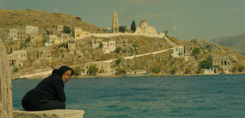 A woman dressed in black seated on a stone bench along the Mediterranean shoreline. There are houses dotted on the rocky hill across the bay from where she sits.