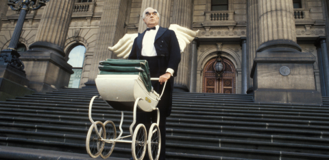 A man in formal evening wear with wings attached to his jacket holds a pram while standing on a steep flight of steps.
