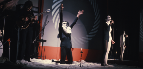 Two mimes in harlequin make-up on a stage with a 1970s rock band.